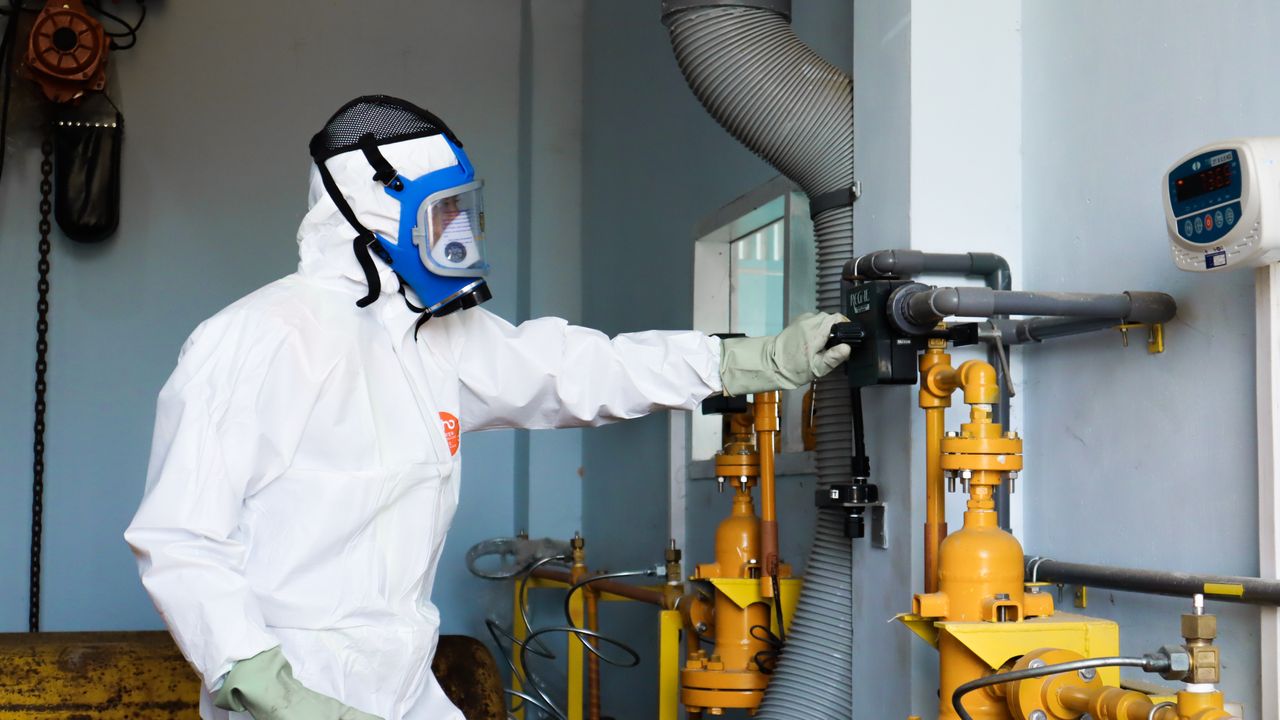 A person in white protective clothing, gloves and a full-face respirator beside industrial pipework.